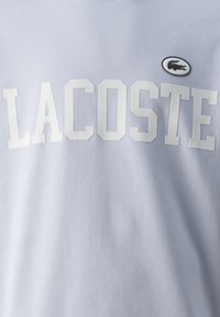 Felpa azzurra chiara con grande testo bianco "LACOSTE" sul petto, accompagnata da un piccolo logo di coccodrillo nero su uno sfondo circolare.
