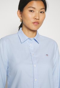 Hele sinine napitärk, mis on valmistatud sileda tekstuuriga kangast. Omab klassikalist kraed ja rinnal väikest broderi logot.