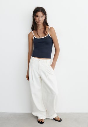 Young woman standing against a white wall wearing a navy camisole, wide white pants, and black flip-flop sandals.
