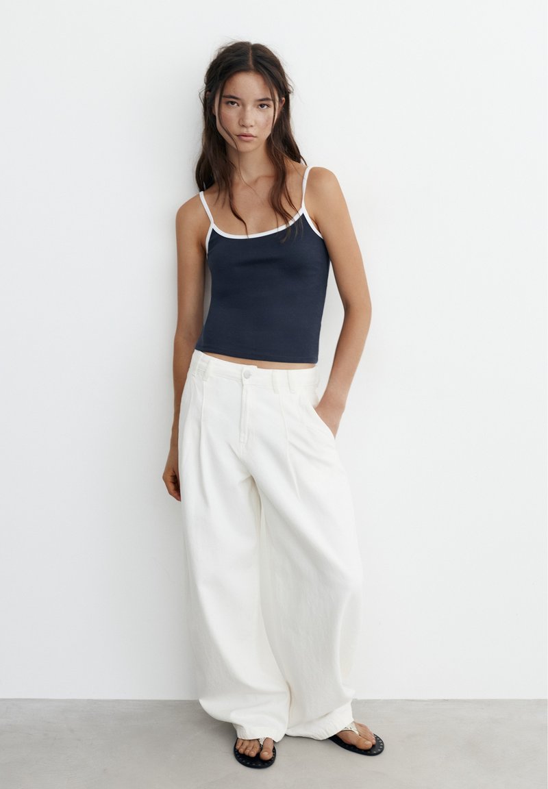 Jeune femme debout contre un mur blanc, portant un caraco bleu marine, un pantalon blanc large et des sandales noires à semelles plates.