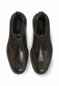 Scarpe eleganti in pelle marrone con finitura lucida, design wingtip, dettagli brogue e chiusura con lacci. Le sole interne nere presentano "HUGO" in rosso.