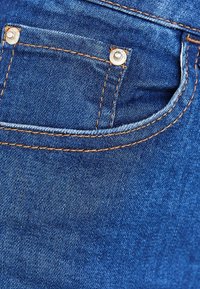 Modrá denimová látka má zadní kapsu s kovovým zapínáním a kontrastním oranžovým stehováním podél okrajů.