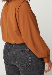 Blusa a maniche lunghe color ruggine con una texture liscia, caratterizzata da pieghe sulle spalle e un polsino con bottoni, abbinata a jeans grigi a vita alta.