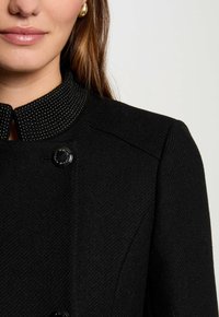 Il cappotto nero presenta un tessuto testurizzato, un colletto a contrasto con piccoli punti argento e chiusure a bottone al collo e alle cuciture delle spalle.
