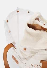 Transparent tote bag med bruna läderhandtag. Innehåller en mjuk, krämfärgad fleeceinsats. Har bruna tryckknappar och logotypdetalj.