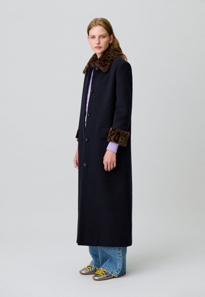 Abrigo largo azul marino con cuello y puños de piel de leopardo, con corte recto, cierre de botones, y llevado sobre una blusa de color púrpura claro.