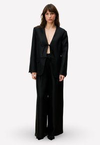 Zwarte blazer en wijde pantalon, gemaakt van gladde stof. De blazer heeft een diepe V-hals en een striksluiting in de taille. Getailleerde pasvorm.