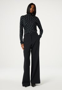 Zwarte lange mouwen top met een klein gouden sterpatroon, gecombineerd met hoge taille wijde broek; model staat met de handen in de zakken.
