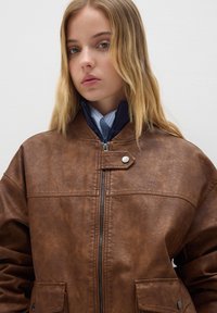 Giacca in finta pelle marrone con chiusura a zip, dettaglio a scatto e due tasche frontali. Presenta un colletto alto e un design sfiancato.