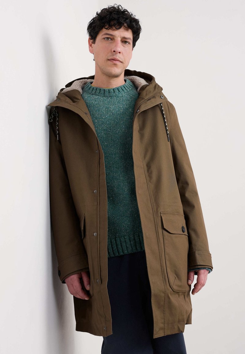 Vyras, atsirėmęs į sieną, dėvi rudos spalvos gobtuvą striukę su pūkuota pamušalu ir žalią mezginį, žvelgia į priekį su neutraliu veido išraiška.