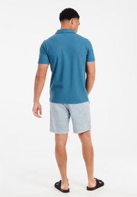 Blauwe polo voor heren, korte mouwen, lichtgewicht stof, gecombineerd met lichte shorts. Zwarte sandalen. Gezien van achteren tegen een witte achtergrond.