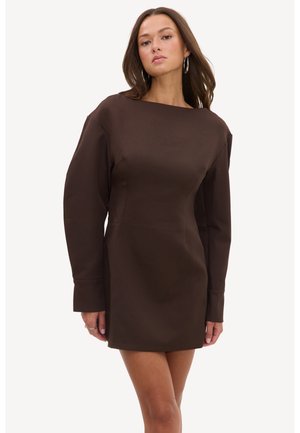 Femme portant une mini-robe marron ajustée à manches longues avec un col montant et des boucles d’oreilles créoles sur un fond uni.