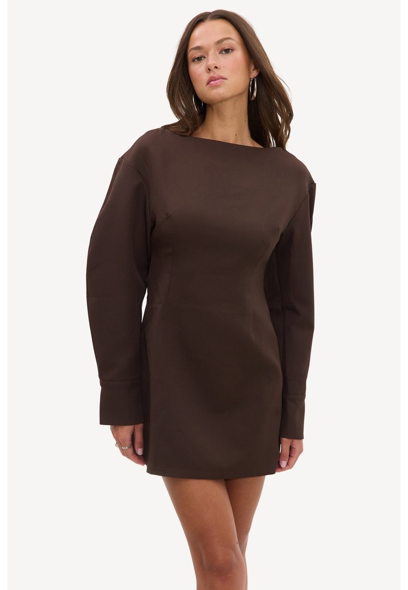 Femme portant une mini-robe marron ajustée à manches longues avec un col montant et des boucles d’oreilles créoles sur un fond uni.