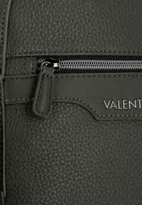 Sac à dos en cuir texturé vert foncé avec une fermeture éclair argentée, des coutures décoratives et un détail de nom de marque embossé à l'avant.