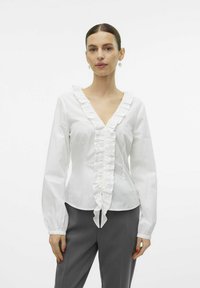 Witte katoenen blouse met een V-hals, voorzien van verticale fronsen aan de voorkant en uitlopende lange mouwen. Gecombineerd met grijze broek.