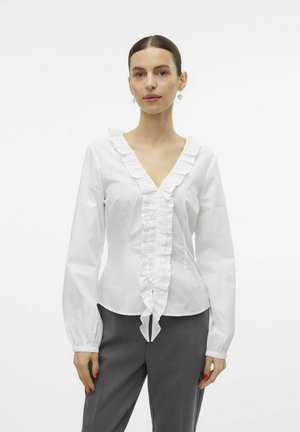 Witte katoenen blouse met een V-hals, voorzien van verticale fronsen aan de voorkant en uitlopende lange mouwen. Gecombineerd met grijze broek.
