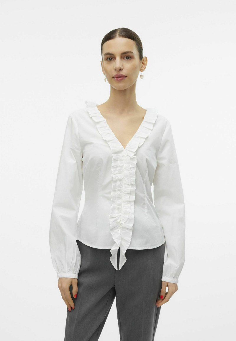 Witte katoenen blouse met een V-hals, voorzien van verticale fronsen aan de voorkant en uitlopende lange mouwen. Gecombineerd met grijze broek.