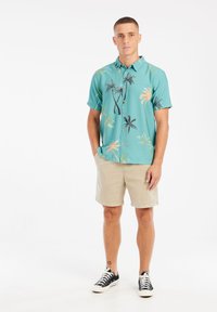 Turquoise shirt met korte mouwen met zwart en pastel palmboomprints, beige shorts en zwarte sneakers met witte veters tegen een witte achtergrond.