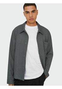 Graues Button-Down-Hemd mit Kragen, Brusttasche und langen Ärmeln, über einem weißen Rundhals-T-Shirt getragen. Glatte Stoffoberfläche.