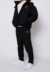 Svart puffjacka med huva och orange logotyp, kombinerad med svarta byxor med logotyptryck och vita sneakers. Slät textur, sportig design.