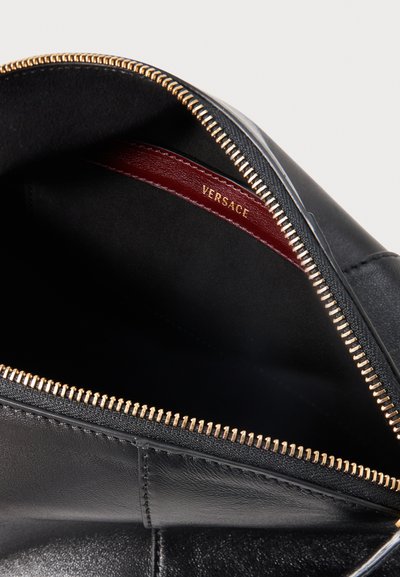 Μαύρη δερμάτινη τσάντα με χρυσλό φερμουάρ μερικώς ανοιχτή, που δείχνει κόκκινη εσωτερική ετικέτα με ανάγλυφο "VERSACE" σε χρυσά γράμματα.