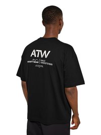 Mężczyzna w czarnej, oversize'owej koszulce, pokazujący tył z białym tekstem graficznym i symbolami, w tym "ATW", "Off Duty Gear" i liczbami.