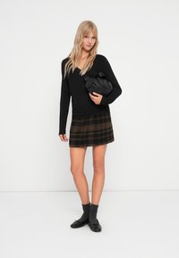 Schwarzes Langarmshirt, brauer karierter A-Linien-Rock, schwarze Umhängetasche, dunkle Socken und schwarze flache Schuhe, vor einem neutralen Hintergrund.