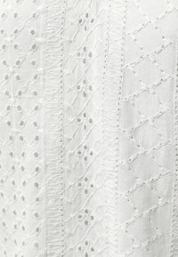 Tissu blanc avec des motifs brodés complexes et de petits œillets disposés en bandes verticales.