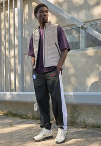 Beige padded vest over a purple t-shirt, paired with black pants featuring a white stripe, and white sneakers with a textured sole.