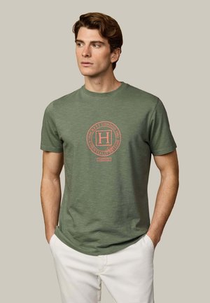 Hombre con camiseta gráfica verde y pantalones blancos, de pie con las manos en los bolsillos, mirando hacia un lado contra un fondo liso.