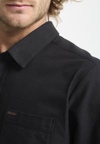 Chemise noire à boutons avec col structuré, comportant une poche plaquée et une petite étiquette de marque. Le tissu est lisse avec une finition mate.
