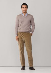 Svetlohnedý sveter s výstrihom do V cez modrú a bielu pruhovanú košeľu, skombinovaný s béžovými kordúrovými nohavicami a tmavohnedými topánkami. Jednoduchý, prispôsobený strih.