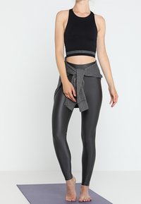 Czarny top na ramiączkach z szarym pasem, połączony z błyszczącymi szarymi legginsami z wysokim stanem. Modelka stoi boso na macie do jogi.