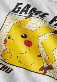 Balts auduma T-krekls ar dzeltenu Pikachu grafiku, melniem kontūriem, sarkaniem vaigiem un ar tekstu "GAME ON" virs režģveida raksta.