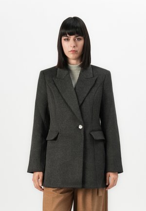 Person med glatt, mørkt hår iført en mørkegrå skreddersydd blazer over en stripete høyhalset topp og brune bukser, som står mot en hvit bakgrunn.