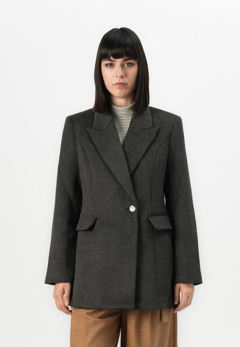 Personne avec des cheveux raides et foncés portant un blazer ajusté gris foncé par-dessus un haut rayé à col montant et un pantalon marron, se tenant devant un fond blanc.