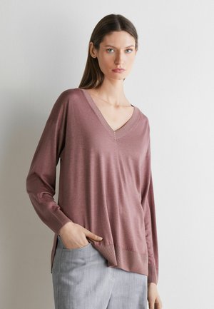 Vrouw met steil bruin haar draagt een stoffig roze V-hals trui en lichtgrijze broek, staand tegen een effen lichte muur.