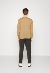Pull beige à manches longues avec texture côtelée, associé à un pantalon sombre et des chaussures blanches. Accent de logo visible près de l'ourlet.