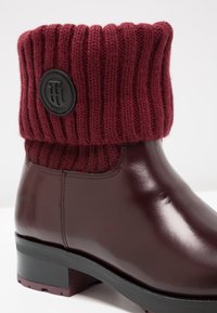 Botte en cuir bordeaux brillant montant à la cheville avec un bord-côtes tricoté assorti et un patch logo circulaire noir.