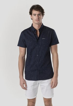 Cămașă bleumarin cu mâneci scurte, cu nasturi și țesătură texturată, având un model cu pete albastru deschis; asortată cu pantaloni scurți albi, suflecați.