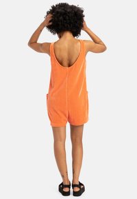 Oranje corduroy romper met een ronde rug, zijzakken en een ontspannen pasvorm, met verticale ribbelstructuur. Zwarte platform sandalen.