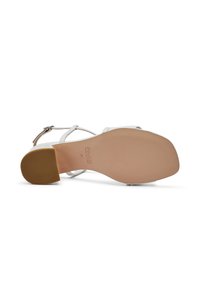 Sandalia blanca con tiras, con un empeine de cuero liso, diseño de punta abierta y suela de goma color bronceado. Cuenta con una correa de tobillo ajustable.