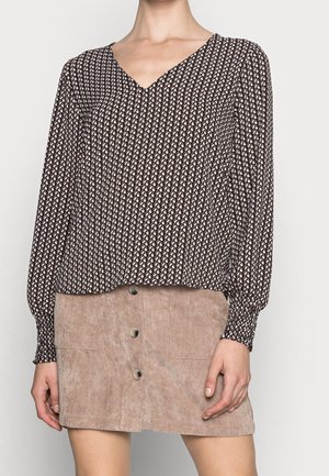 Woman wearing a brown patterned long-sleeve V-neck blouse and a beige buttoned corduroy skirt, standing against a light background.
