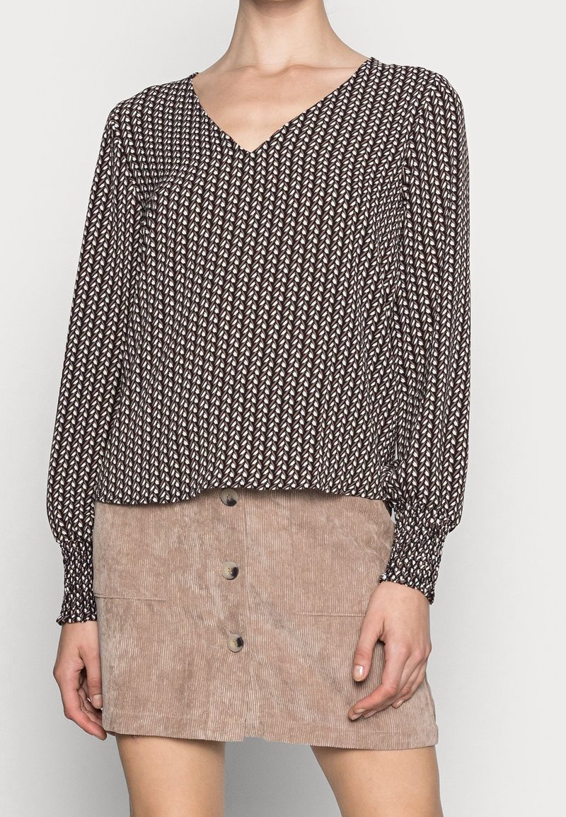 Femme portant un chemisier marron à manches longues à motifs et col en V, et une jupe en velours côtelé beige boutonnée, debout devant un fond clair.