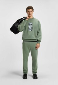 Felpa verde oliva con stampa grafica e logo, abbinata a pantaloni jogger coordinati. Incluse scarpe da ginnastica nere e una borsa a tracolla nera.