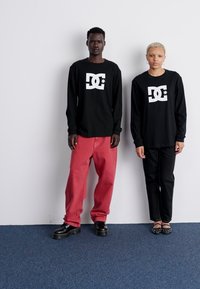 Sorte langærmede skjorter med hvidt logo, røde baggy jeans og sorte bukser. Sorte sko med tykke såler; modellen poserer foran en ensfarvet baggrund.