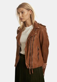 Braune Leder-Motorradjacke mit asymmetrischen Reißverschlüssen, Druckknopfkragen und zwei Reißverschlusstaschen. Gestylt über einem weißen T-Shirt und einem plissierten Rock.