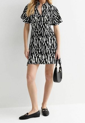 Woman wearing black and white animal print short dress, holding black handbag, and wearing black flat loafers.