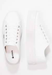 Baskets en toile blanches avec une semelle texturée, bout rond, design à lacets et rayures latérales discrètes. Présentent le logo "victoria" sur la semelle intérieure.