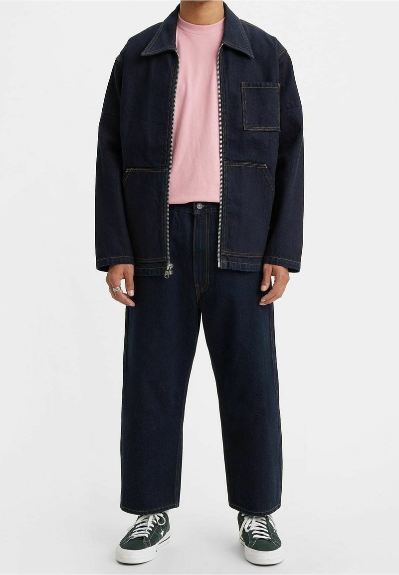 Mörk denimbomulljacka med dragkedja fram, bröstficka och avslappnad passform, tillsammans med en ljus rosa t-shirt och mörka vida jeans. Gröna sneakers.